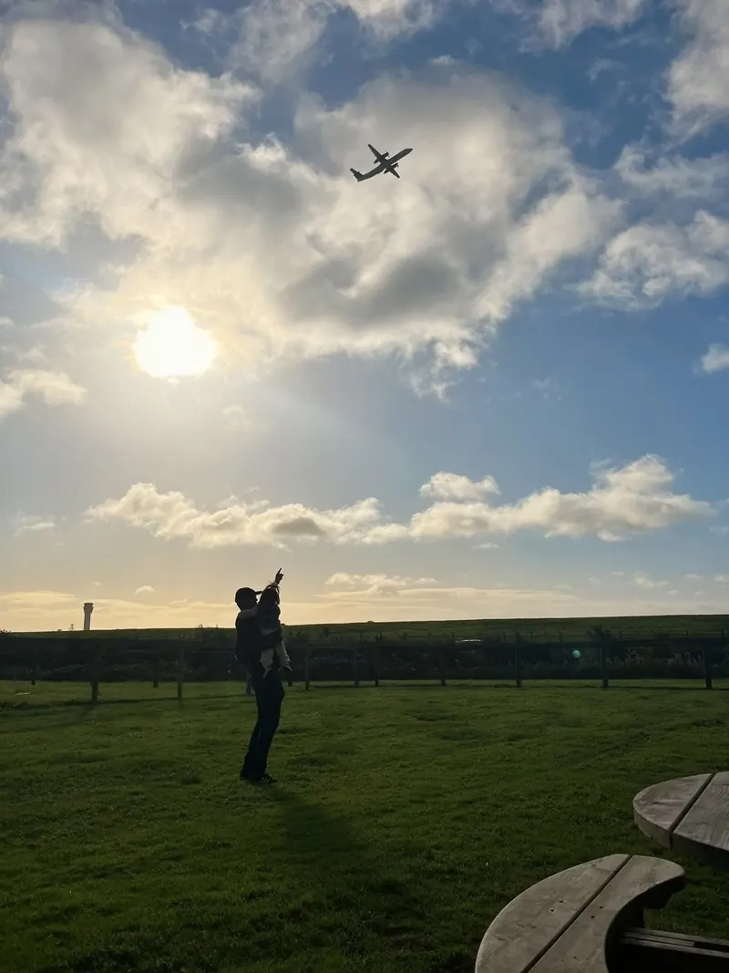 A dad holding his young daughter on his hip, both in silhouette against a bright evening sky, as she points up at a small plane flying overhead.